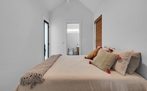 Ein Schlafzimmer mit Bett, Kissen, einer Tagesdecke, einem Fenster mit Bambusrollvorhang und Blick ins Bad.
