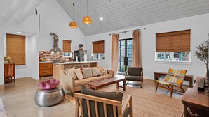 A spacious living area with a sofa, coffee table, and chairs, a kitchen with a brick backsplash and modern appliances, and large windows with blinds. - DogTrot Hotel Suite A (Waco)
