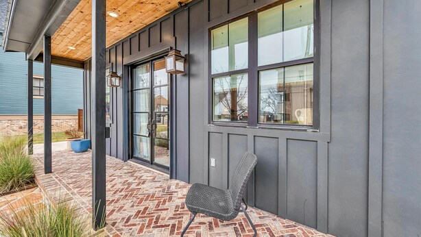 A covered patio with a brick floor, a chair, and a building with large windows and a dark exterior. - DogTrot Hotel Suite A (Waco)