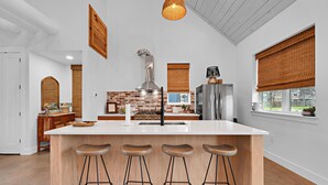 A modern kitchen with a large island, wooden stools, a brick backsplash, and a stainless steel range hood. - DogTrot Hotel Suite A (Waco)