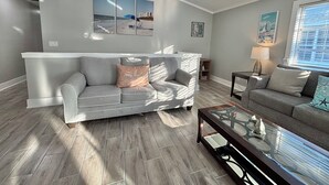 A living room with a grey sofa, a glass coffee table, and a small side table with a lamp. There are wall-mounted pictures and a window with blinds.