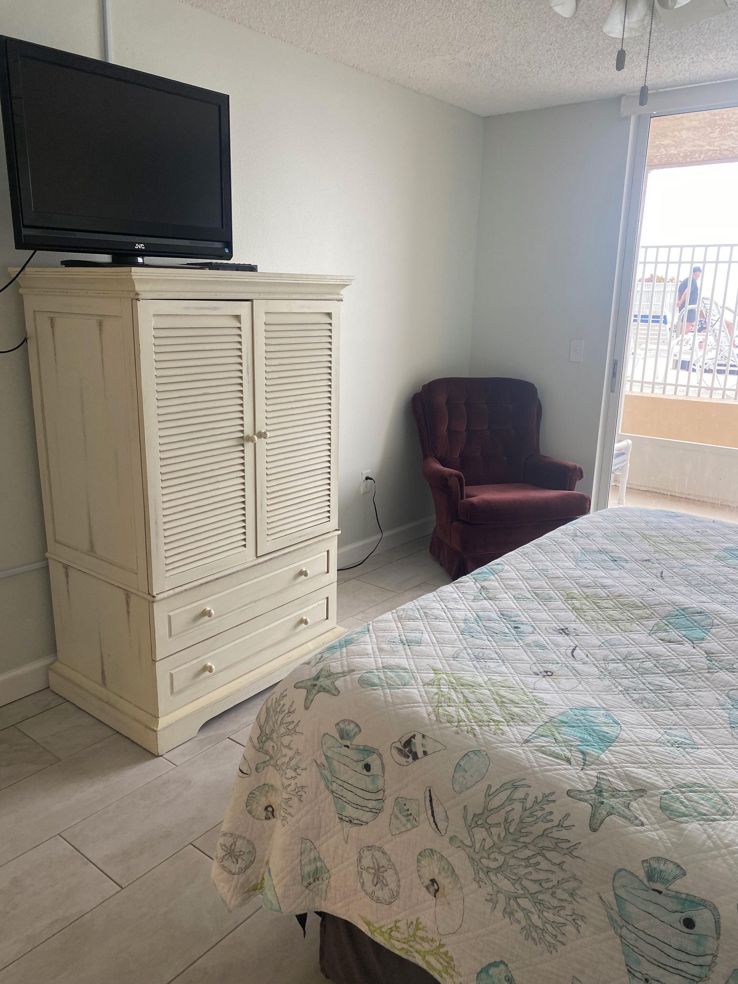 Un dormitorio con una cama, un televisor en un mueble, una silla y vistas al exterior.