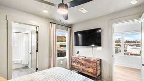 A bedroom with a bed, a ceiling fan, a TV mounted on the wall, and a view of the outside through a window.