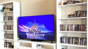 Ett vardagsrum med en platt-TV som visar logotypen för ”Walt Disney Pictures” på skärmen, omgivet av bokhyllor fyllda med DVD-filmer och videospel.