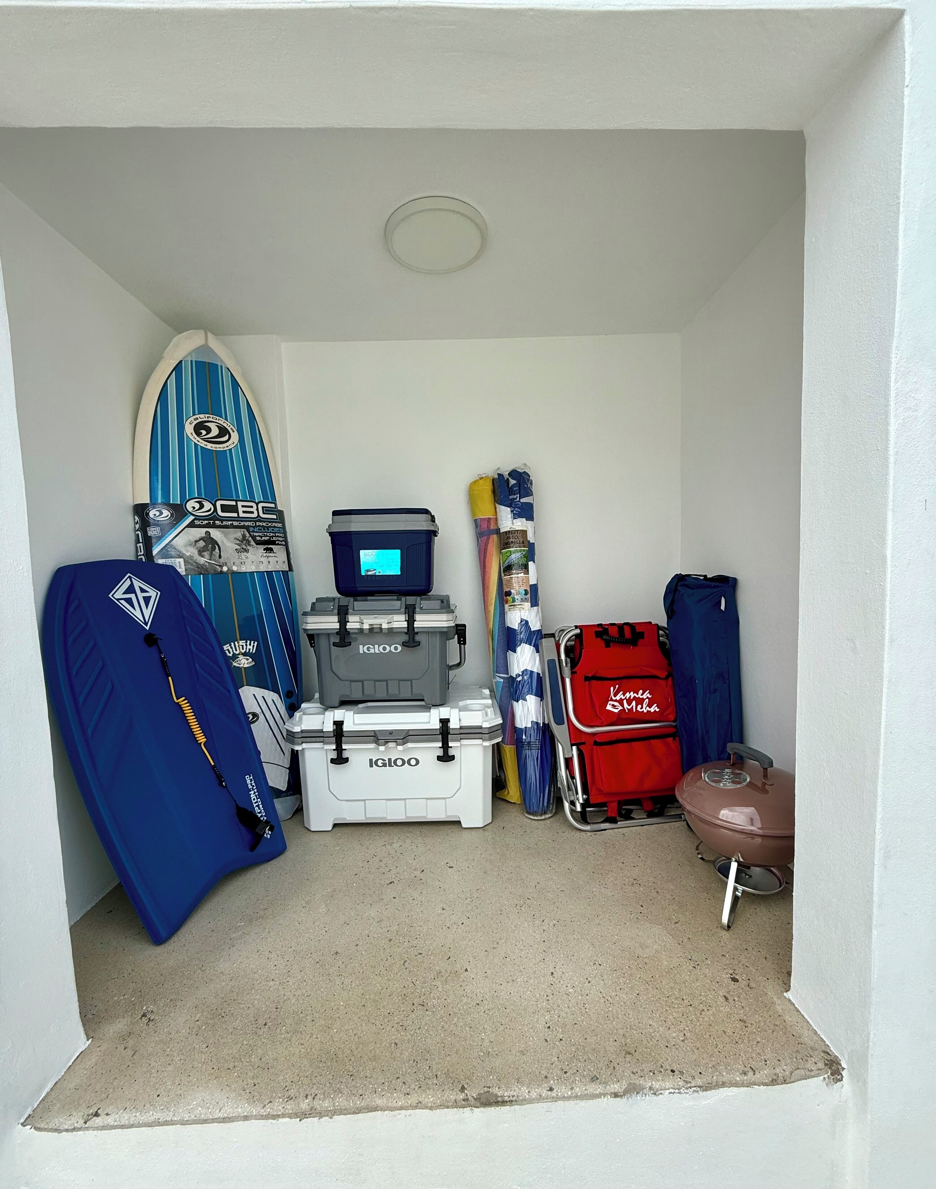 A small room with a surfboard, a suitcase, a red bag, and a storage box.