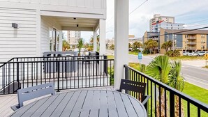 A balcony with a table and chairs, overlooking a street with buildings and palm trees. - 50 Steps To Beach, Heated Pool, Hot Tub, Gulf View, Dog Friendly, Lost Dunes 4BR (Gulf Shores)