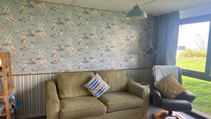 A living room with a green sofa, a black chair, a patterned wallpaper, and a view of the outdoors. - Cosy Holiday Park Chalet, Quiet Location, Child Friendly, Near Bude (Penstowe Holiday Park, Kilkhampton)
