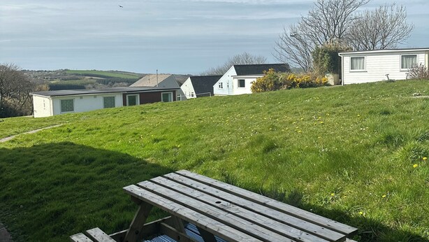 A picnic table on a blue deck with a grassy area and houses in the background. - Cosy Holiday Park Chalet, Quiet Location, Child Friendly, Near Bude (Penstowe Holiday Park, Kilkhampton)