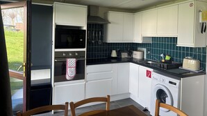 A compact kitchen with white cabinets, a microwave, oven, and a washing machine. - Cosy Holiday Park Chalet, Quiet Location, Child Friendly, Near Bude (Penstowe Holiday Park, Kilkhampton)