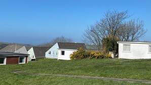 A row of single-story houses with a grassy lawn in front, a clear blue sky, and hills in the background. - Cosy Holiday Park Chalet, Quiet Location, Child Friendly, Near Bude (Penstowe Holiday Park, Kilkhampton)