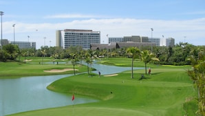 Ein Golfplatz mit einem Wasserhindernis, Palmen und einem großen Gebäude im Hintergrund.