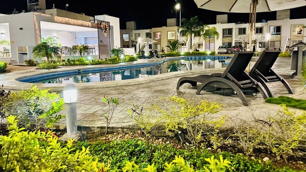 A nighttime pool area with lounge chairs and umbrellas, surrounded by greenery and modern buildings. - Playa del Carmen Apartment (Playa del Carmen)