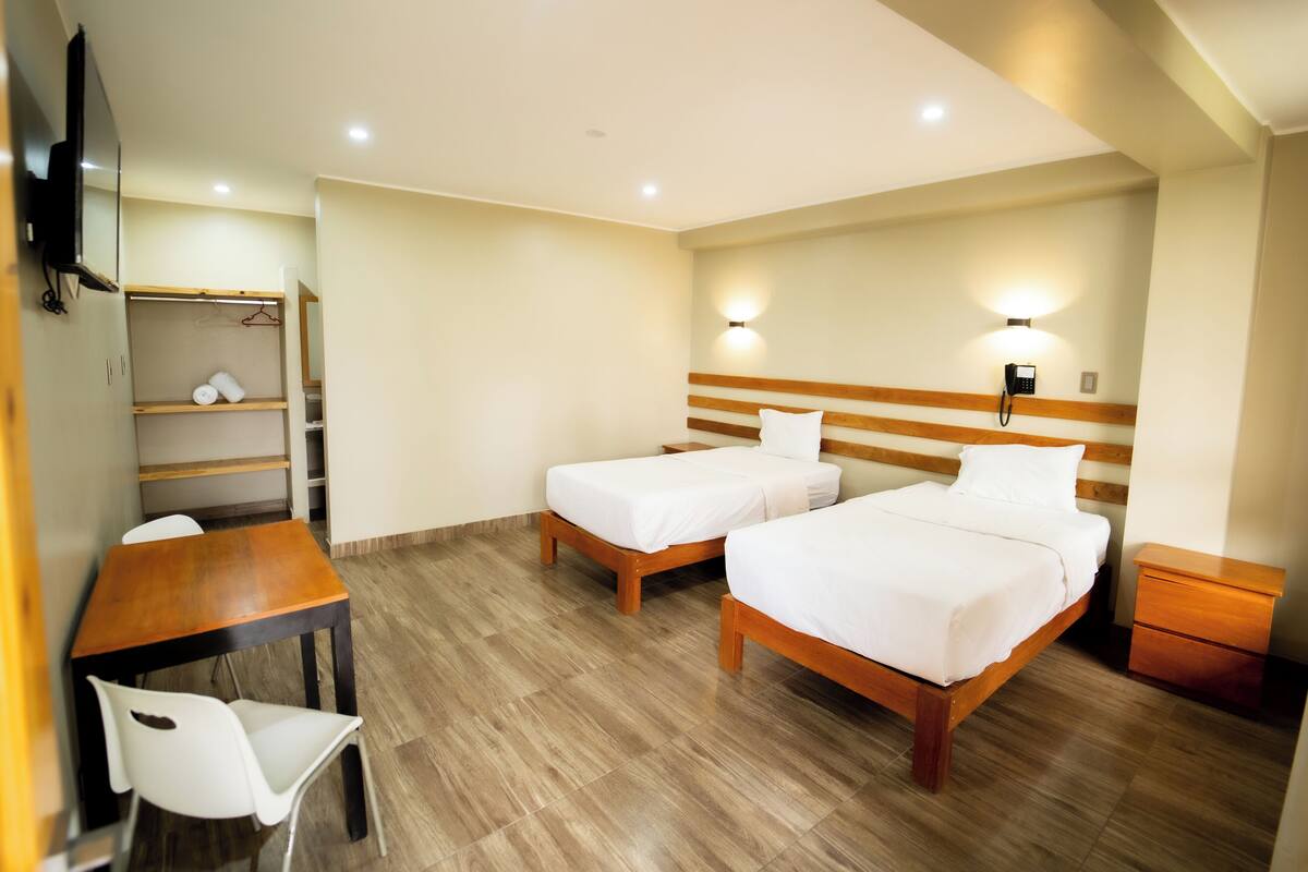 A hotel room with two beds, a wooden desk, a chair, a wardrobe, and a bedside table.