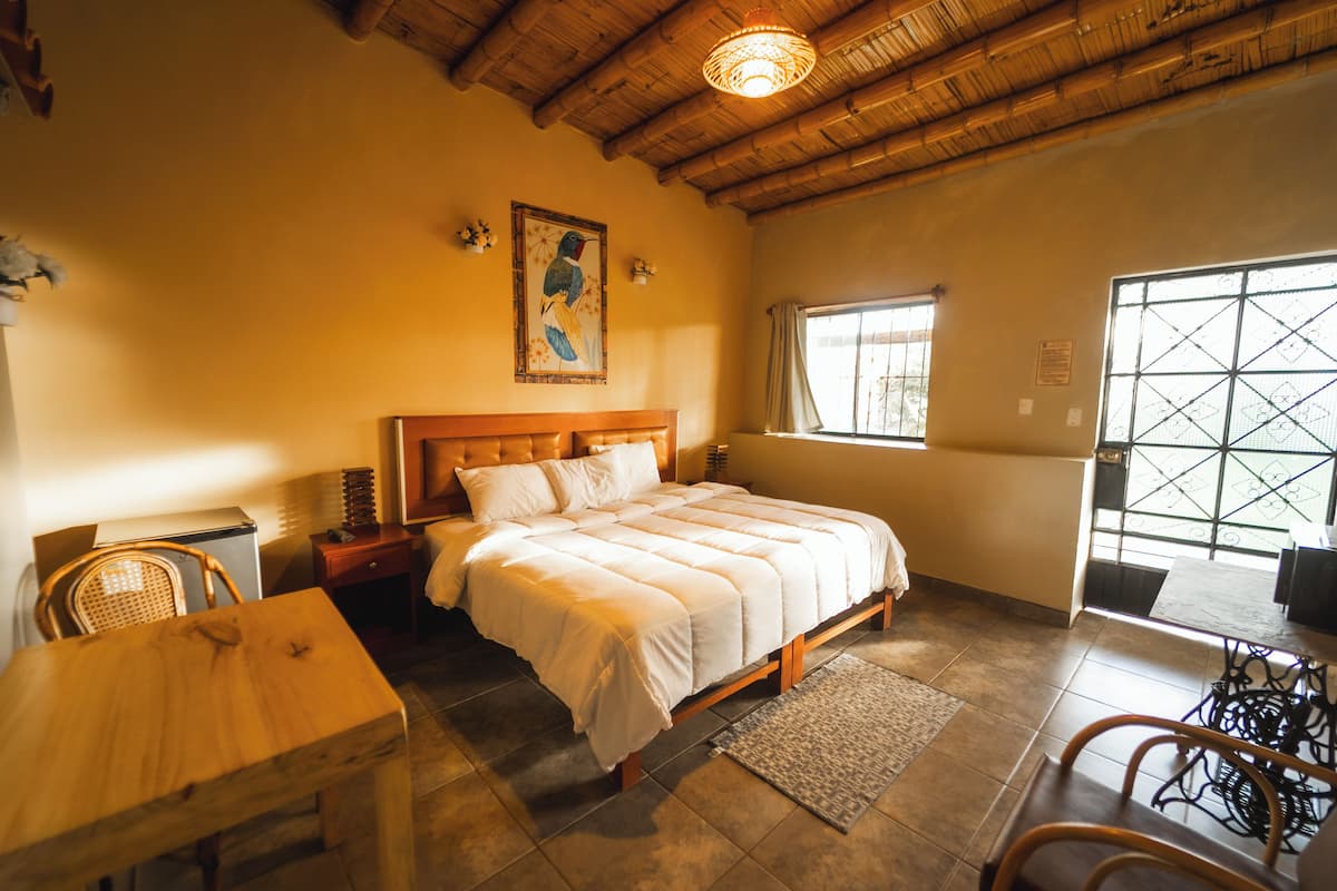 A bedroom with a wooden bed, a dining table, and chairs. There is a window with a view of the outside.