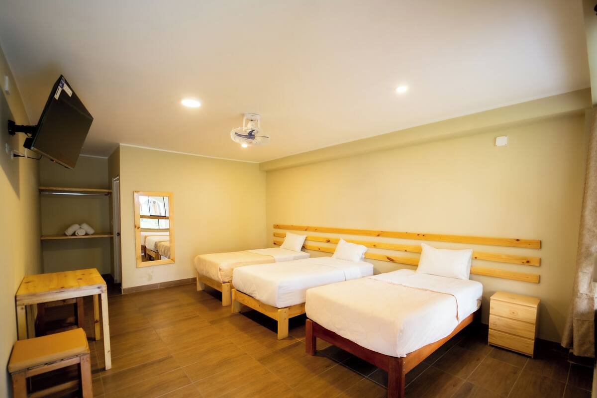 A room with two beds, a wooden table, a small stool, a shelf with towels, and a mounted TV.