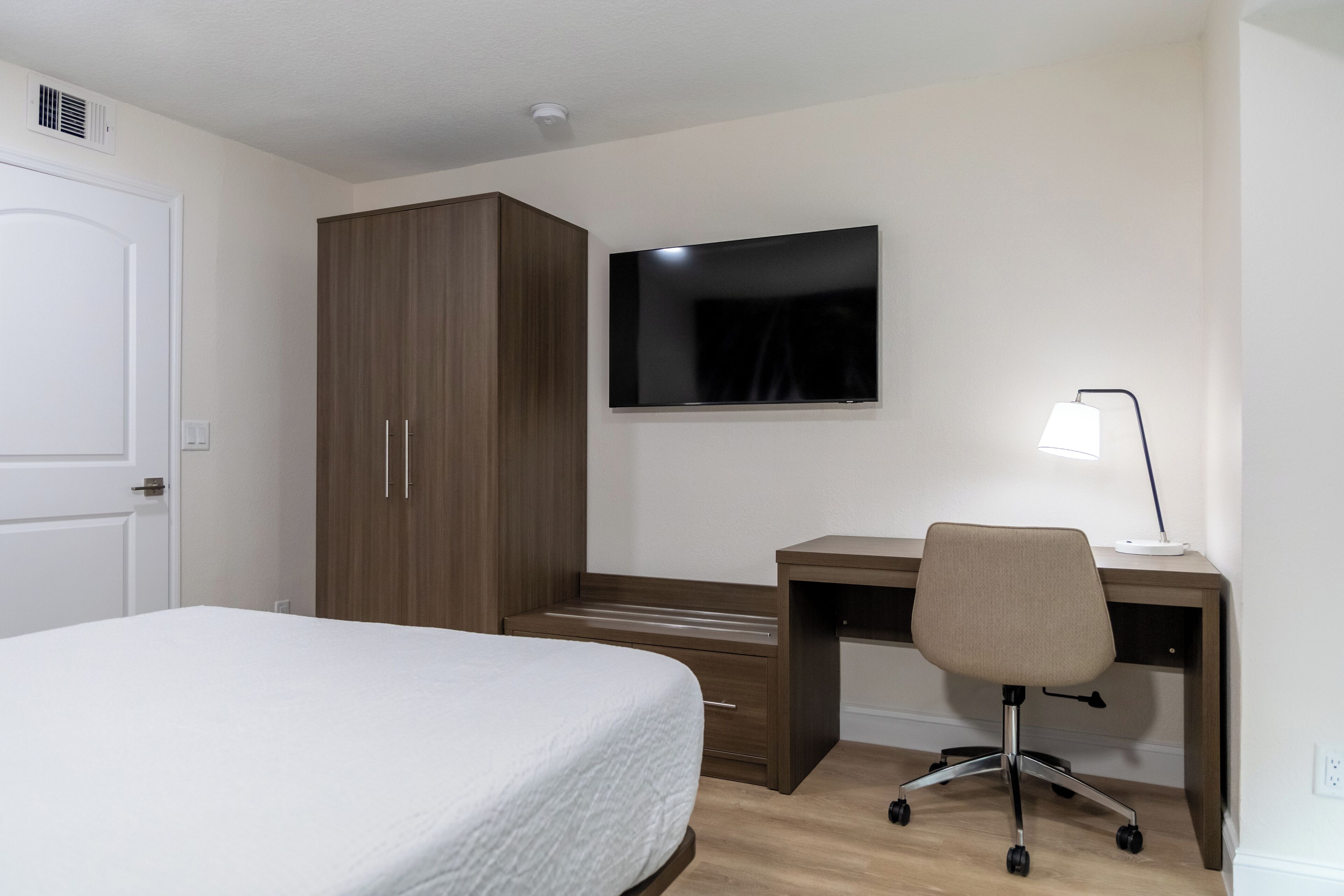 Un dormitorio con cama, armario, escritorio con silla y televisor de pantalla plana.