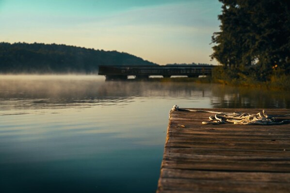 A wooden dock extending into calm water with a rope on it, a bridge in the background, and a forested hill.