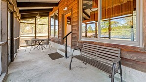 A covered porch with a wooden bench and metal table, large windows, and a view of trees.