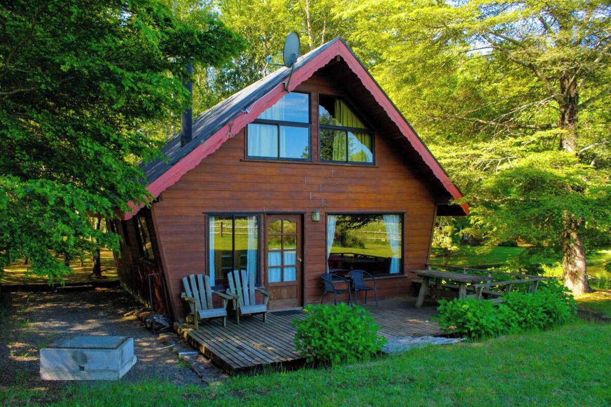 Une cabane en bois dotée d’un toit en pente, de grandes fenêtres et d’une terrasse avec des sièges d’extérieur.