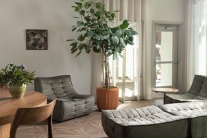 Uma sala de estar moderna com uma mesa redonda de madeira, dois sofás cinzas estofados, uma grande planta em vaso e uma pequena mesa redonda.