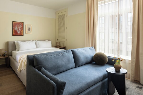 Una habitación de hotel con cama, sofá, una mesita con un jarrón de flores y ventana con cortinas.