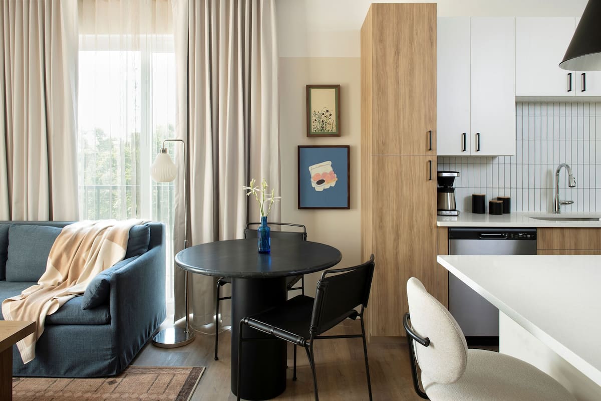 A modern living room with a sofa, a dining table with chairs, a kitchen area with cabinets, and a window with curtains.
