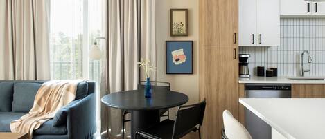 A modern living room with a sofa, a dining table with chairs, a kitchen area with cabinets, and a window with curtains.