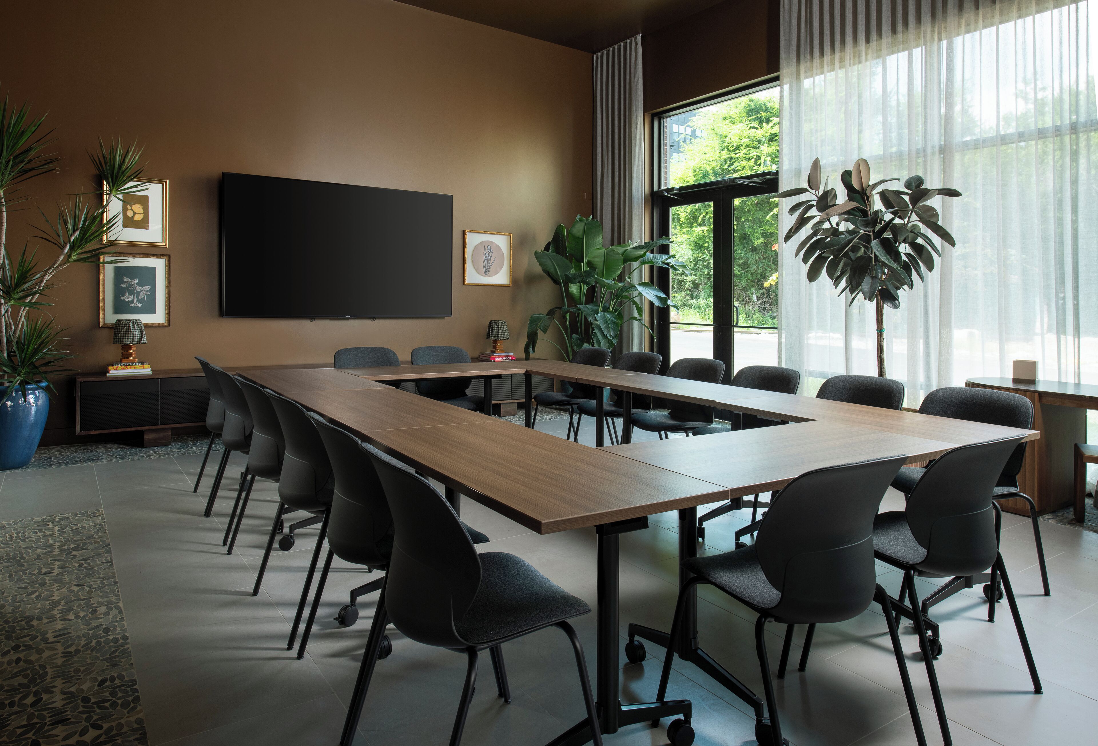 Un salón de conferencias con una larga mesa, sillas, una pantalla de televisión y plantas.