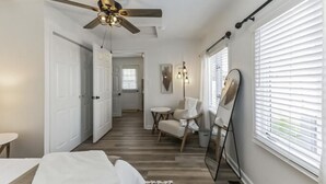 A bedroom with a bed, a ceiling fan, a chair, a table, and a window with blinds.