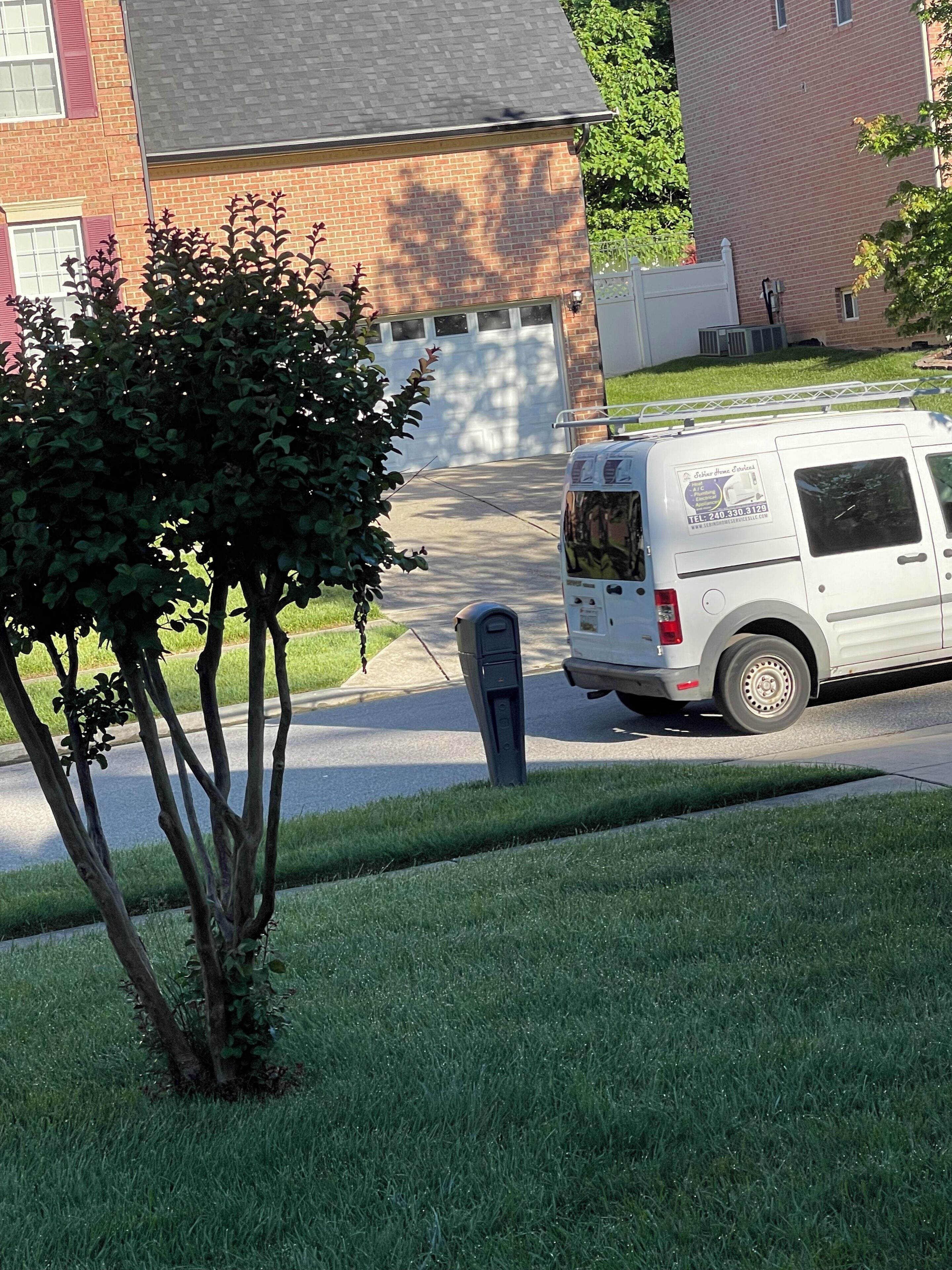 A white van parked on a residential street with a mailbox and a small tree in the foreground.