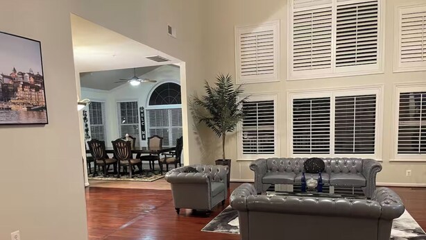 A spacious living room with a large grey sofa, a dining area with a table and chairs, and a large window with blinds. - Lex Holmes Luxury House (Fort Washington)