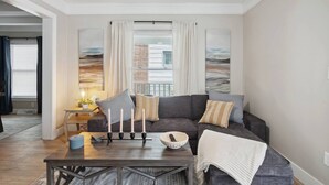 Ein modernes Wohnzimmer mit einem grauen Sofa, einem Holztisch und Blick auf die Stadt durch große Fenster mit weißen Vorhängen.