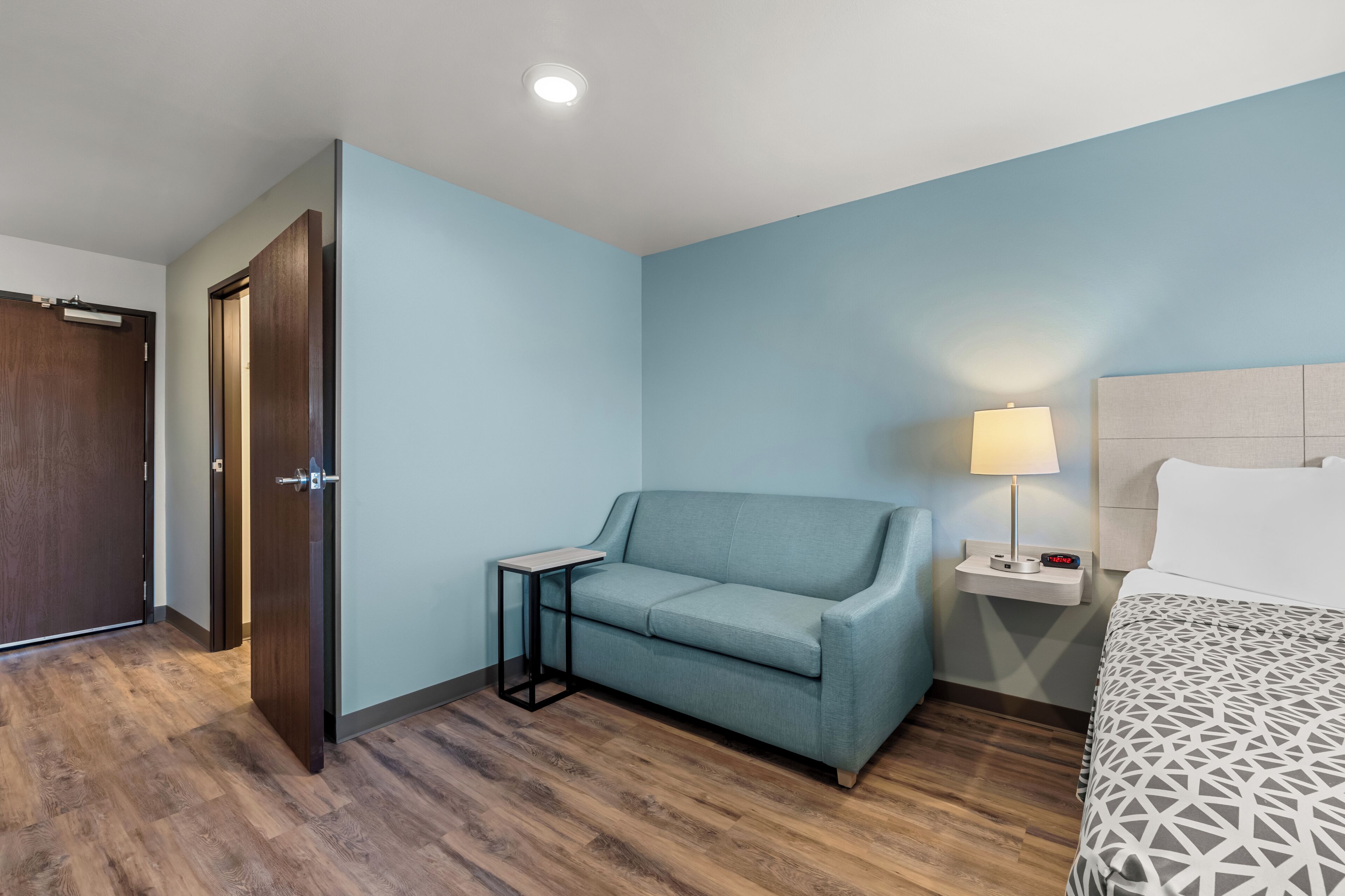 Habitación de hotel con piso de madera, pared azul claro, un sofá, una mesita de noche con lámpara y una cama con colcha estampada.