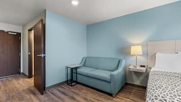 Habitación de hotel con piso de madera, pared azul claro, un sofá, una mesita de noche con lámpara y una cama con colcha estampada.