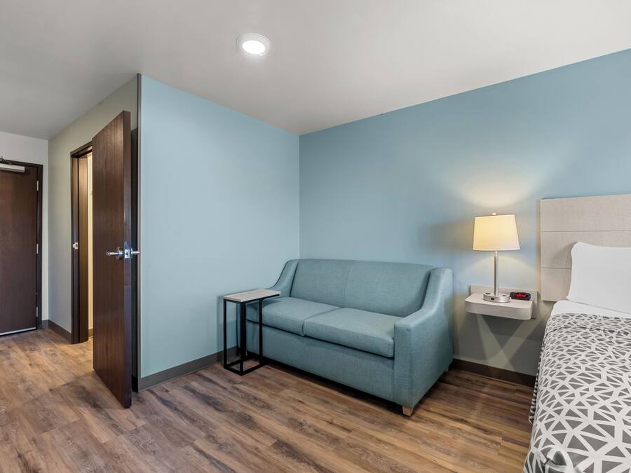 Habitación de hotel con piso de madera, pared azul claro, un sofá, una mesita de noche con lámpara y una cama con colcha estampada.