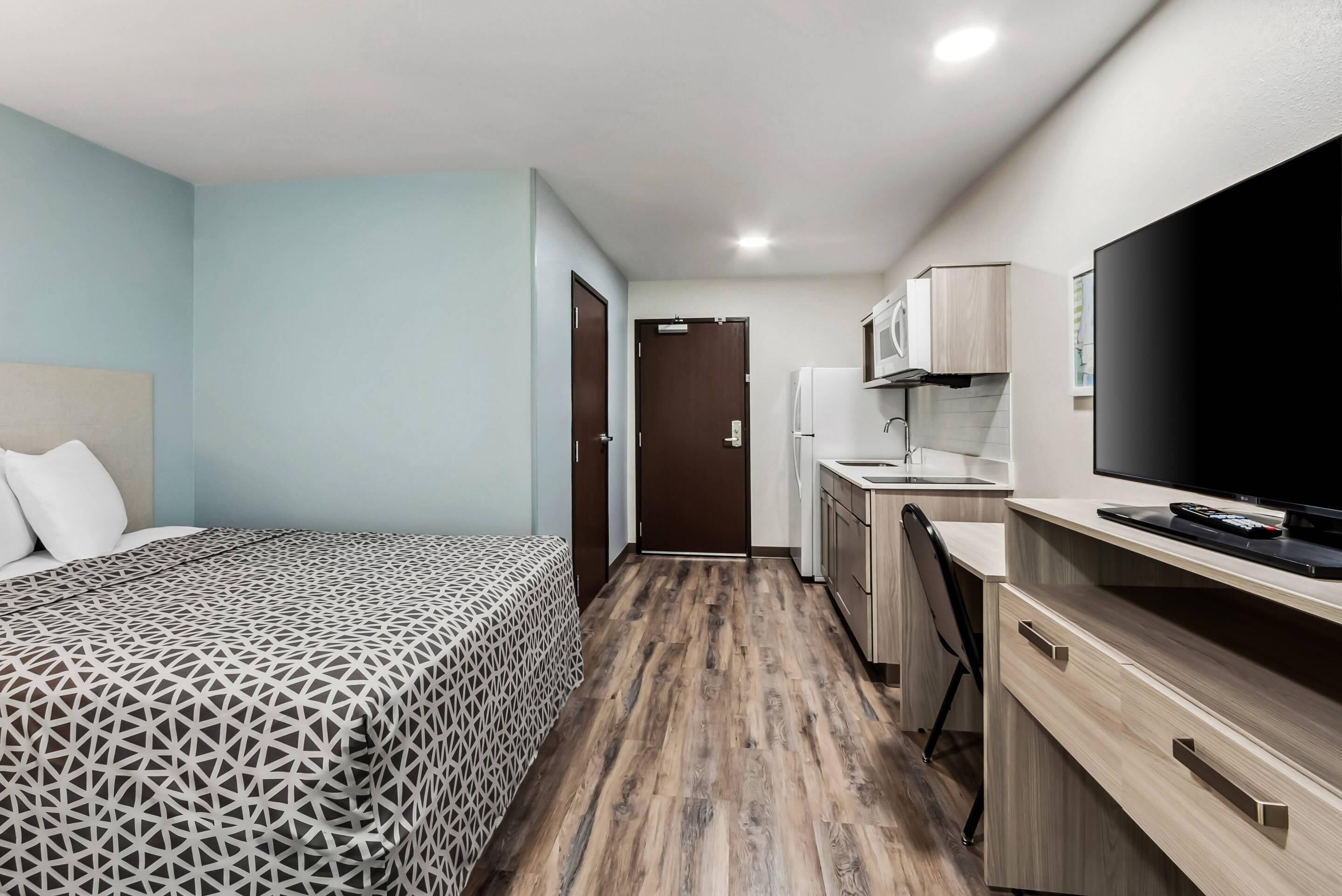 A compact hotel room with a bed, a desk, a chair, a television, and a kitchenette.