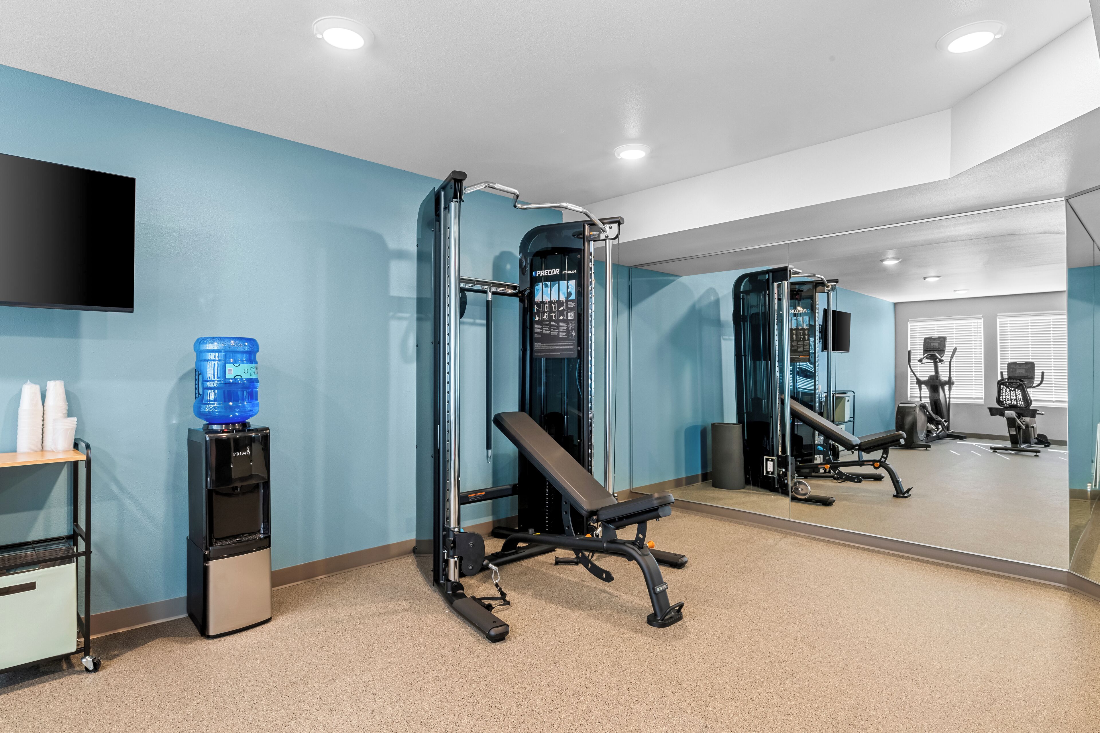 A gym with a bench, weight machine, water dispenser, and a mirror reflecting the equipment.
