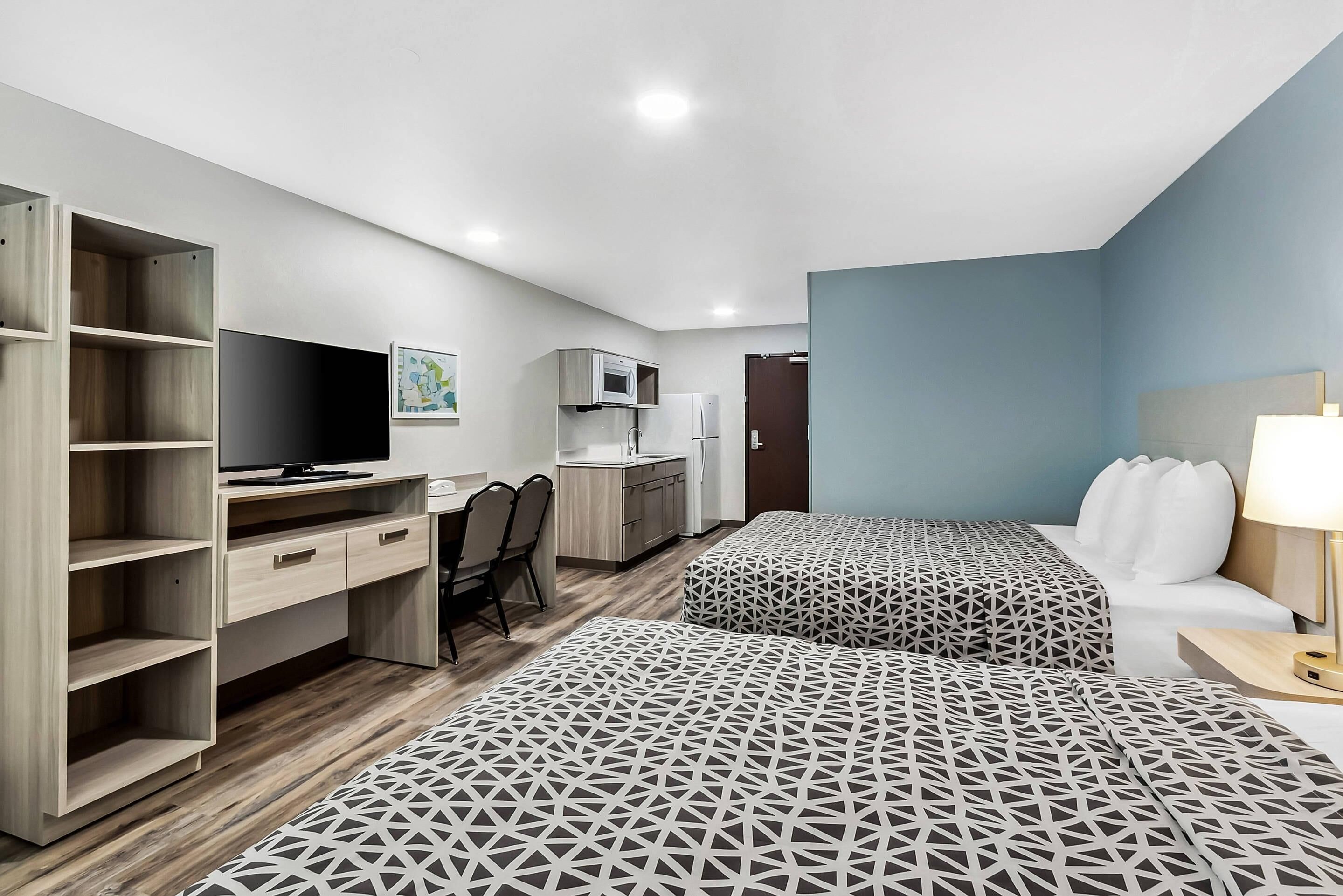 A modern hotel room with a desk, two beds, a TV, a microwave, and a kitchenette.