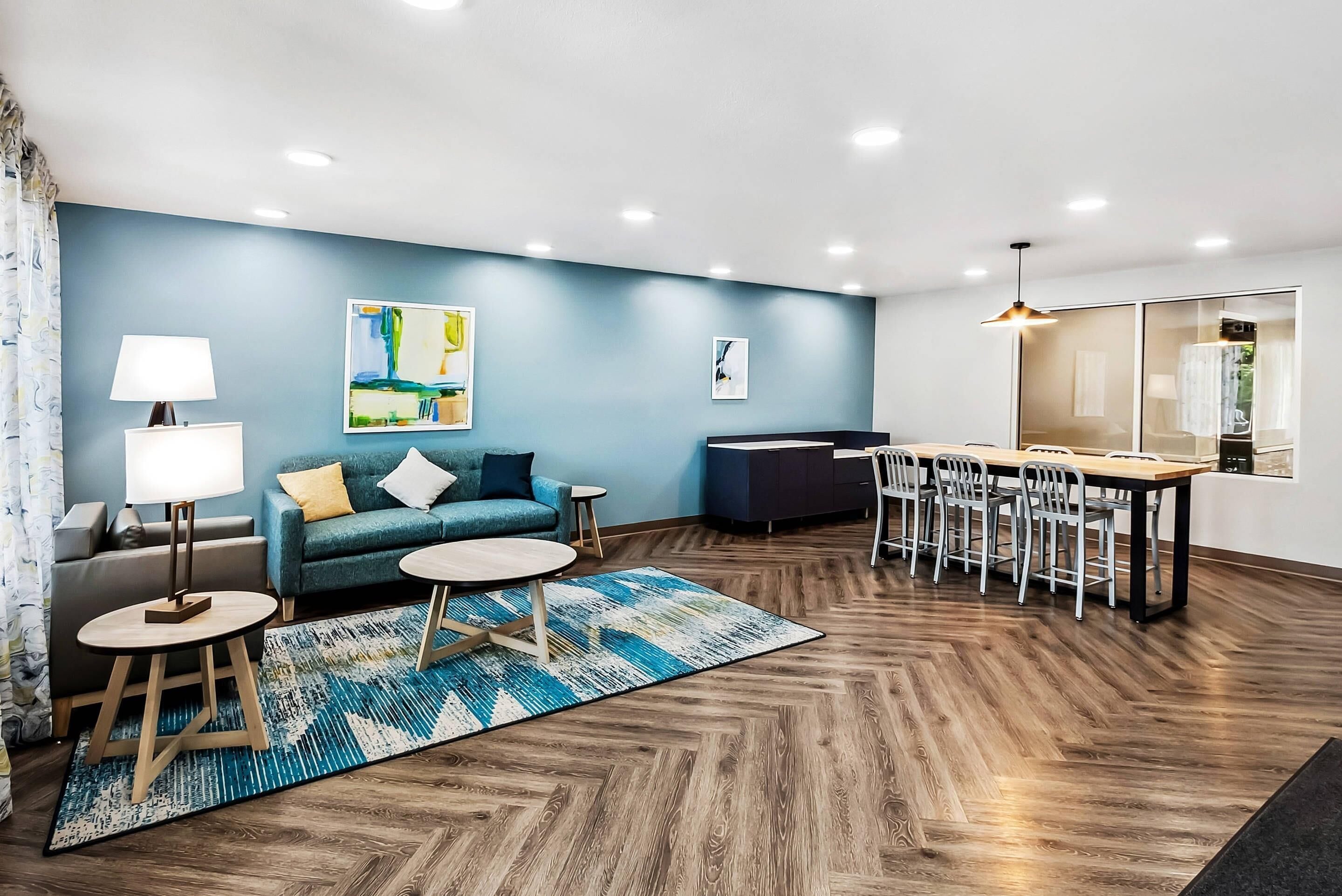 A modern living room with a blue accent wall, a sofa, a coffee table, a dining table with chairs, and a patterned rug.