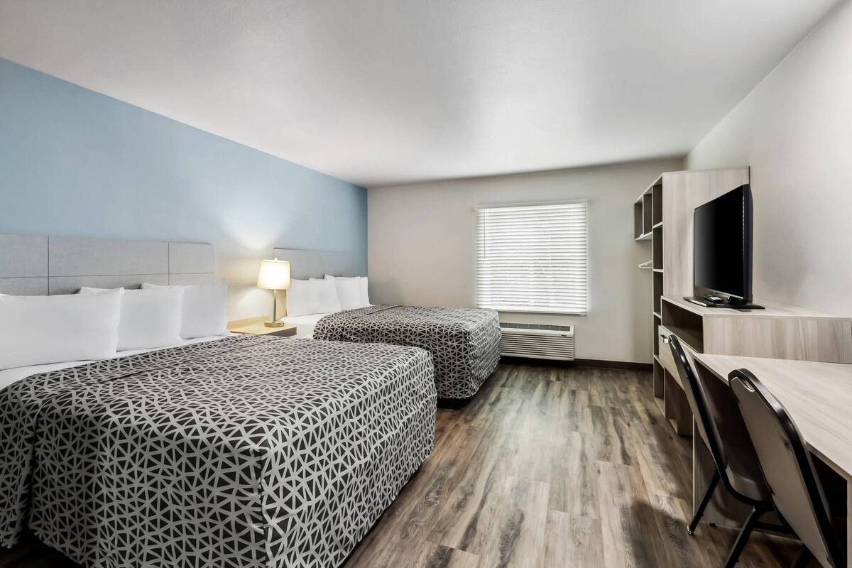 A hotel room with two beds, a desk, a TV, and a window with blinds.