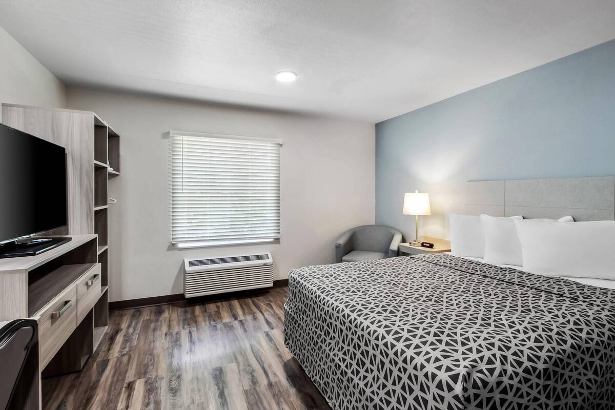 A hotel room with a bed, a desk with a TV, a chair, a bedside table with a lamp, and a window with blinds.