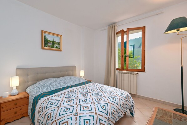 A bedroom with a bed, a nightstand, a lamp, a picture on the wall, a window with curtains, and a radiator.