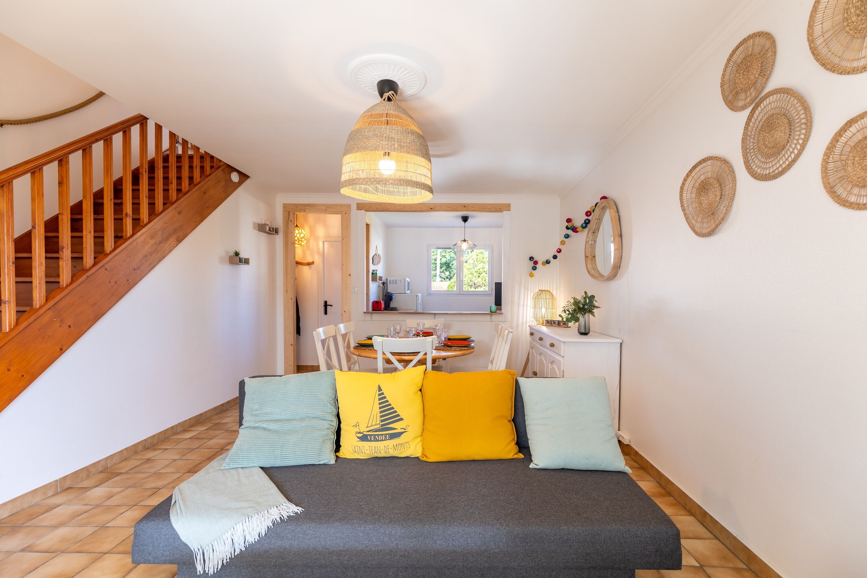 A cozy bedroom with a wooden staircase, a bed with colorful pillows, a woven ceiling light, and a dining area in the background.
