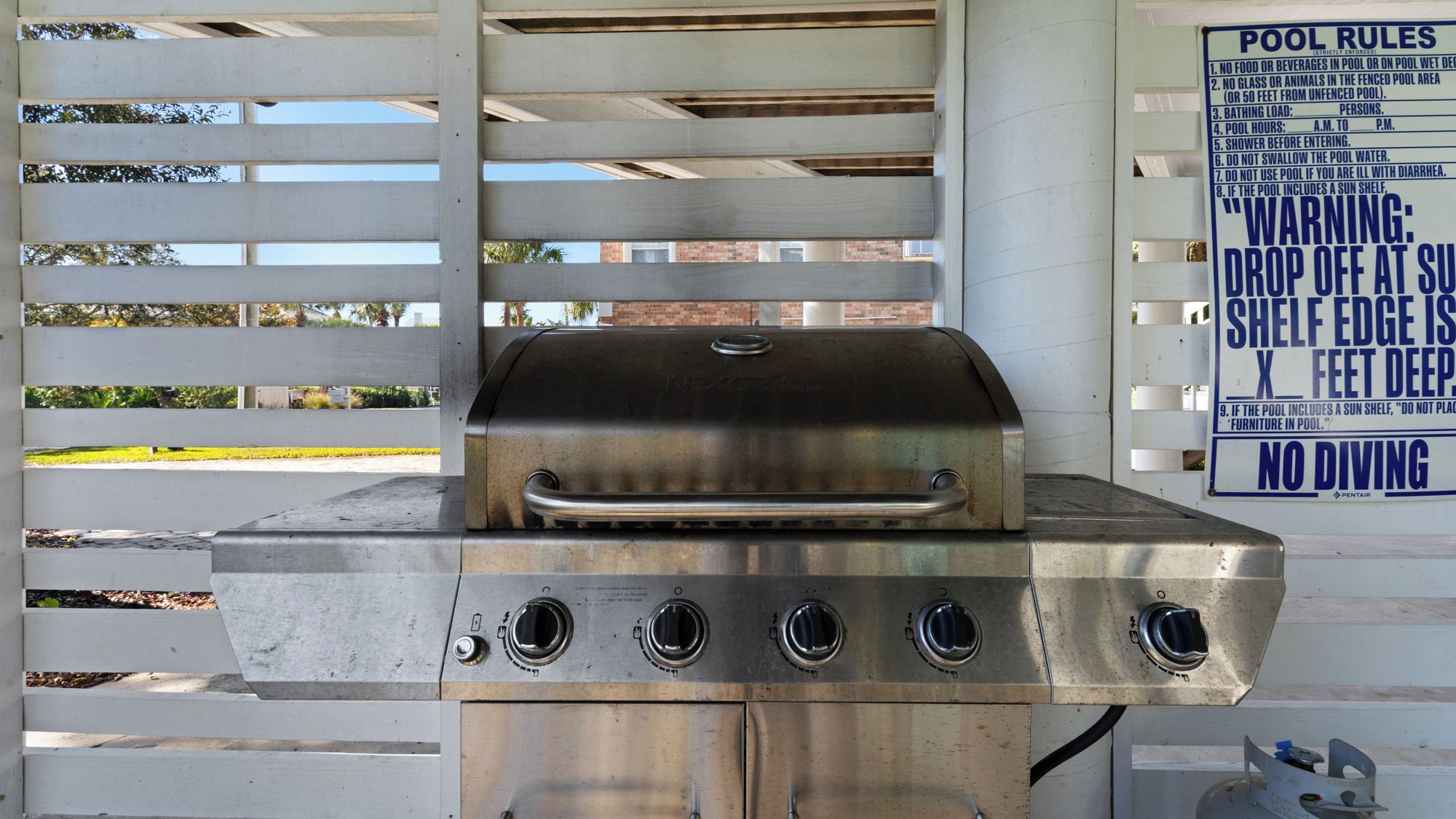 Un barbecue en acier inoxydable doté de quatre brûleurs, d’une étagère pour se protéger du soleil et d’un panneau indiquant les règles à respecter au bord de la piscine.