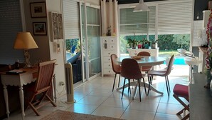 A spacious living area with a dining table and chairs, a sofa, and a large window with blinds. - Charming house with pool and garden, 10 minutes from the sea (Biscarrosse)