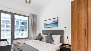 A modern bedroom with a large bed, a wooden wardrobe, a nightstand with a lamp, and a city view through the window. - Bloomfields Executive 1BR Oasis Masdar (Abu Dhabi)