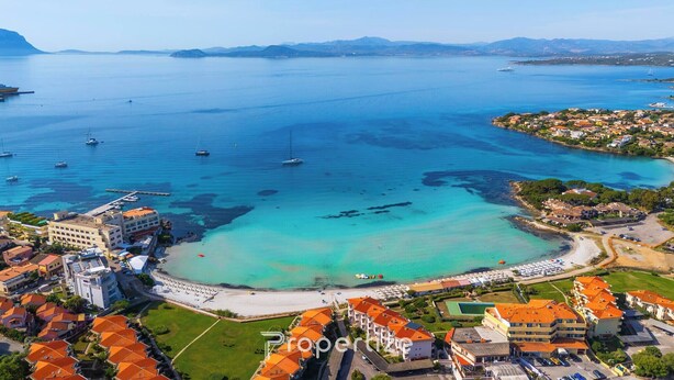 Vue aérienne d’une ville côtière avec un port de plaisance, des eaux d’un bleu limpide et une plage parsemée de parasols.