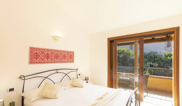 A bedroom with a bed, a bedside table, a lamp, a wall-mounted light, and a sliding glass door leading to an outdoor balcony with plants.