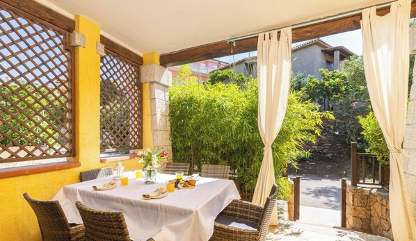 A dining area with a table set for two, surrounded by wicker chairs, a yellow wall, and a view of greenery and buildings in the background.
