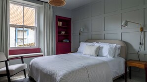 A bedroom with a bed, a chair, a window with curtains, a red bookshelf, and a wall-mounted lamp. - Ledger's Cottage- Harbourfront Home Pittenweem (Pittenweem)